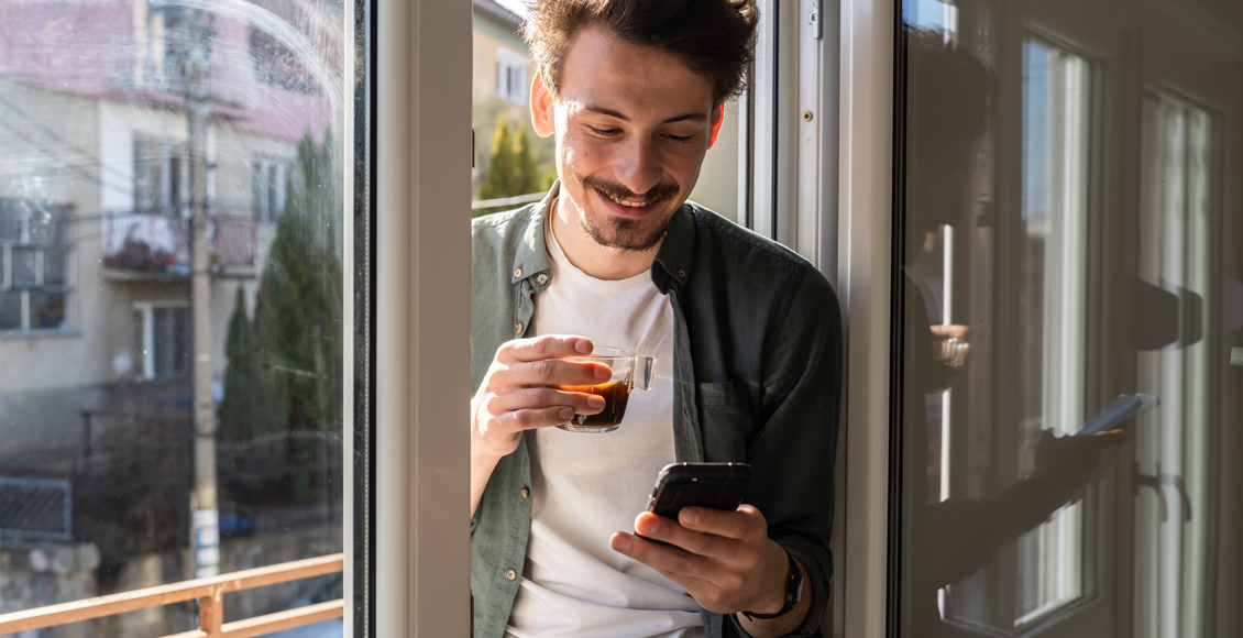 Mann sitzt am offenen Fenster und hält lächelnd einen Kaffeebecher während er auf sein Smartphone schaut.